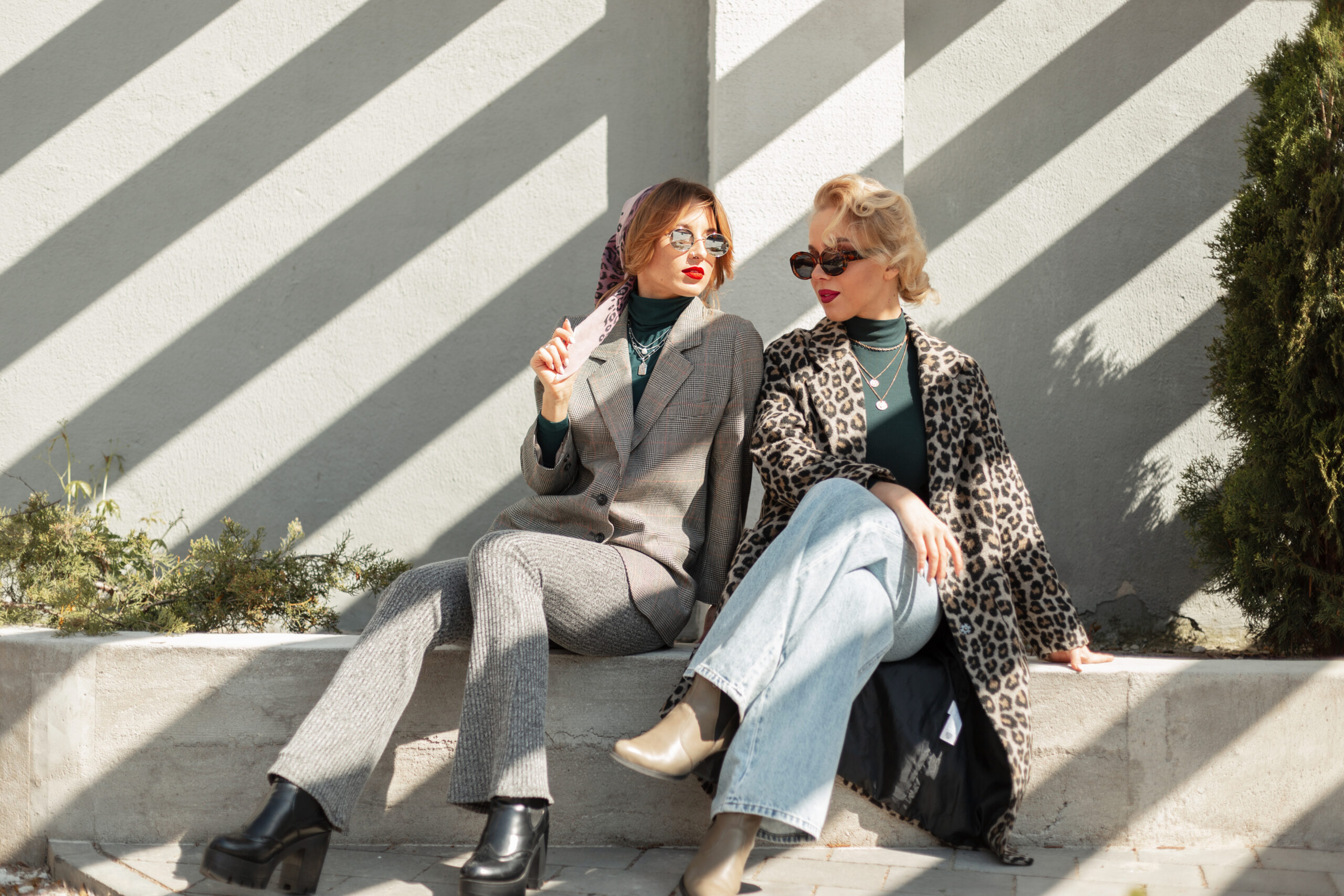 Two beautiful stylish girlfriends of the model in fashionable clothes with leopard coats, clush jeans, a suit and shoes sit in the city with sunlight. Urban vintage style and fashion outfit
